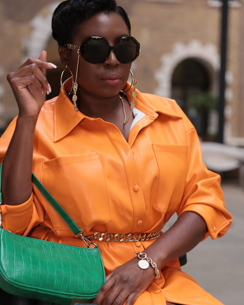 A lady standing wearing a orange dress and a green bag